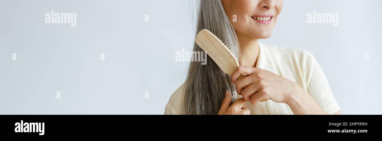 Donna asiatica premurosa di mezza età spazzolare i capelli grigi posando su sfondo chiaro Foto Stock