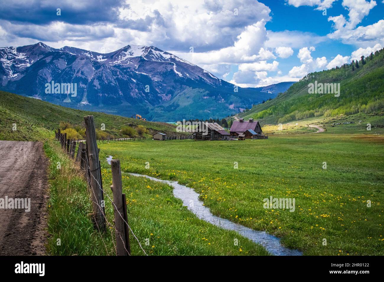 Scena alpina con strada sterrata fienile rustico e ruscello di neve che scorre attraverso la valle con fiori selvatici - montagne innevate vicino a Crested Butte Colo Foto Stock