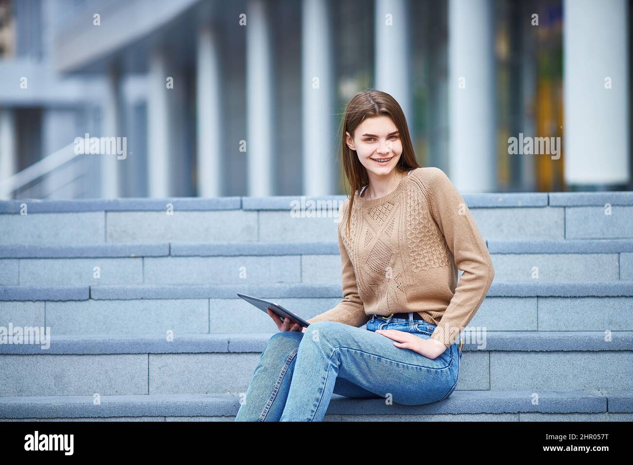 la giovane ragazza si siede sui gradini della strada con un tablet nelle mani e guarda la macchina fotografica sorridendo. Spazio di copia. Foto Stock