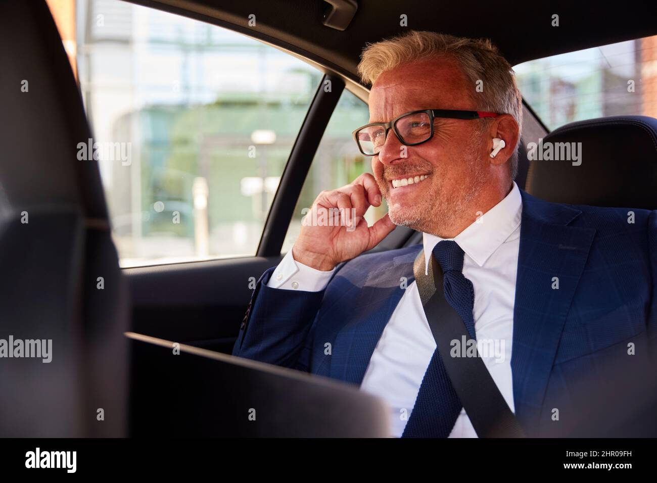 Uomo d'affari maturo che indossa auricolari wireless che lavorano su un notebook in taxi o in auto Foto Stock