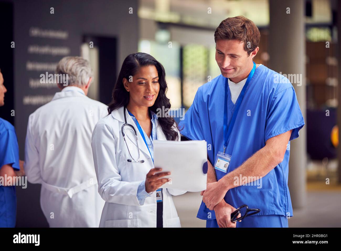 Due medici in cappotti bianchi e scrub con tavoletta digitale che hanno informale riunione in ospedale Foto Stock