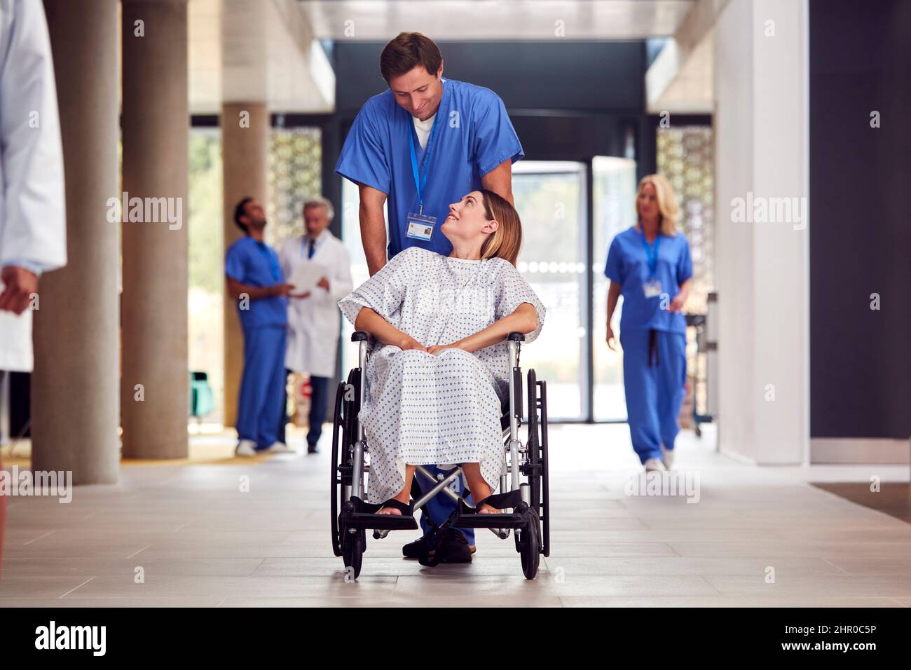 Infermiera maschile che indossa scrub spingendo il paziente femminile in sedia a rotelle attraverso l'edificio dell'ospedale Foto Stock