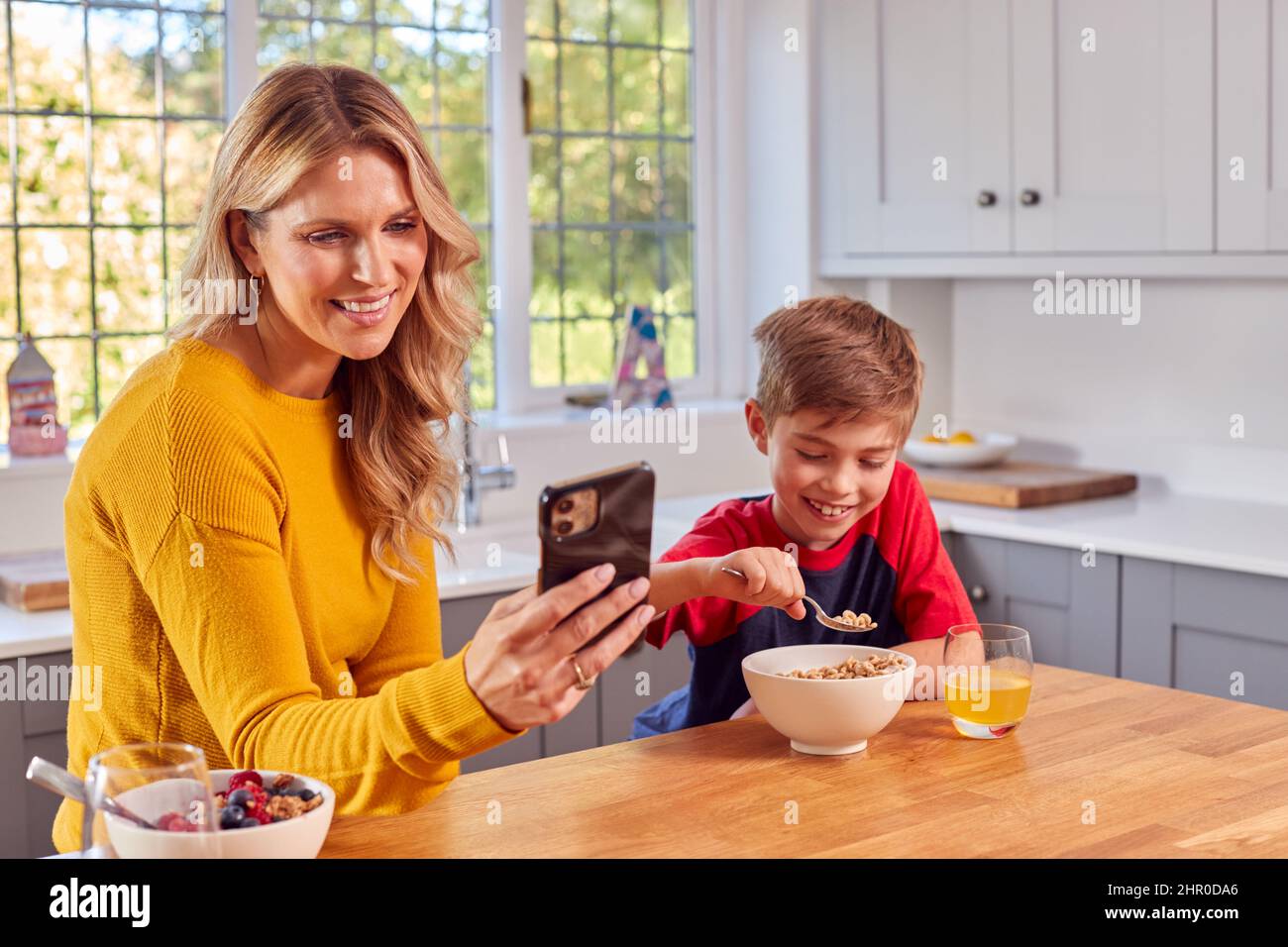Il figlio a casa che mangia la colazione cereale al banco della cucina mentre la madre guarda il telefono mobile Foto Stock