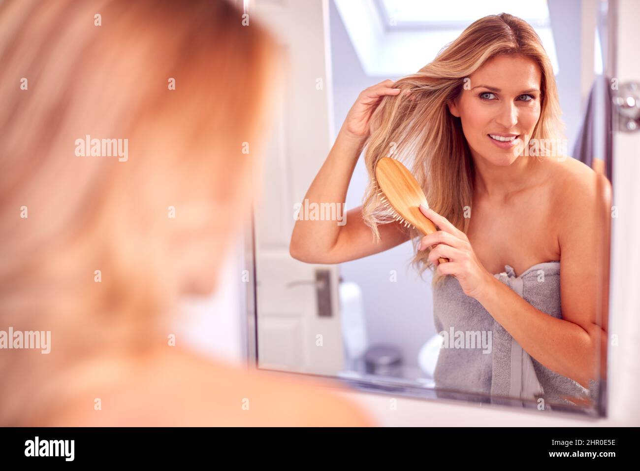 Donna matura che prende i capelli spazzolando guardando nello specchio in bagno a casa Foto Stock
