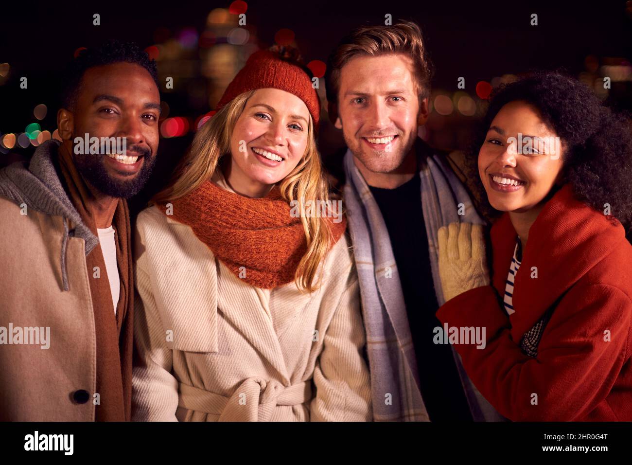 Ritratto di amici all'aperto indossando cappotti e sciarpe incontro contro le luci della città di notte Foto Stock