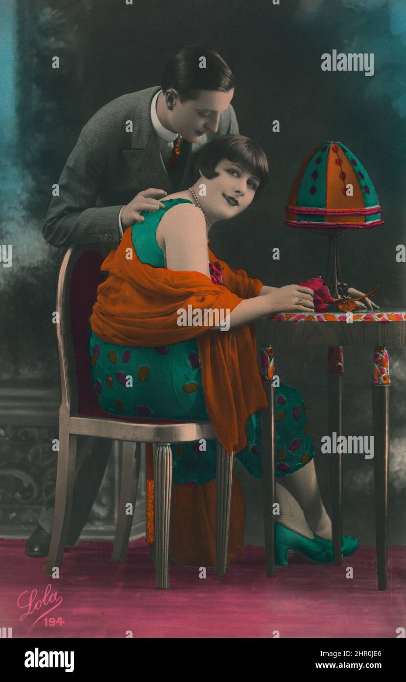 Foto dipinta in stile retrò. Una donna si siede su una sedia a un tavolo e guarda la macchina fotografica mentre un uomo si appoggia dolcemente su di lei. Foto Stock