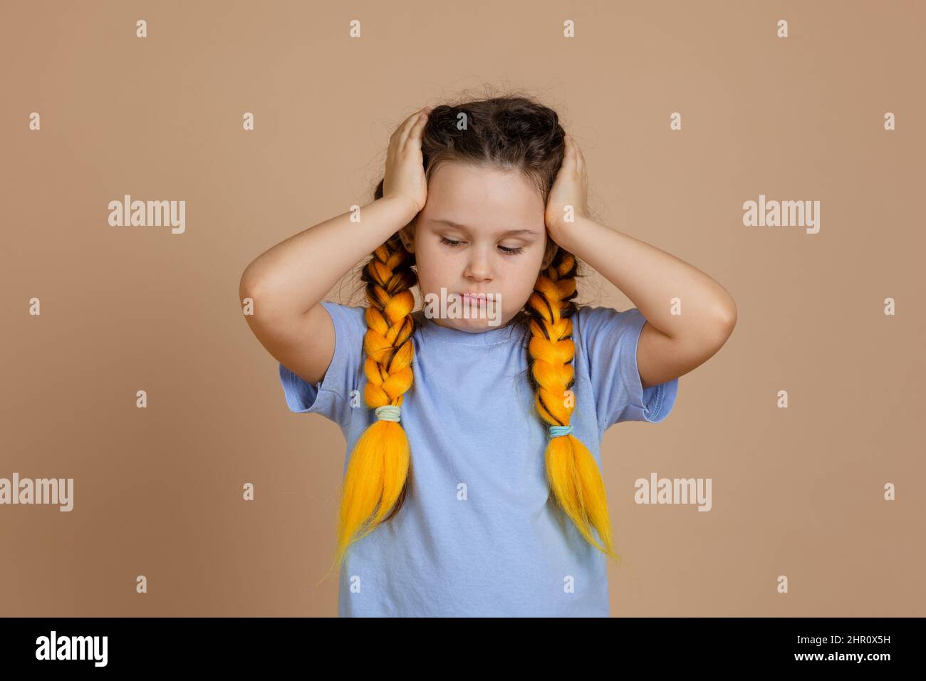 Arrabbiato caucasico bambino femmina che tiene testa con le mani guardando verso il basso avendo kanekalon trecce di colore giallo sulla testa in blu t-shirt sul beige Foto Stock