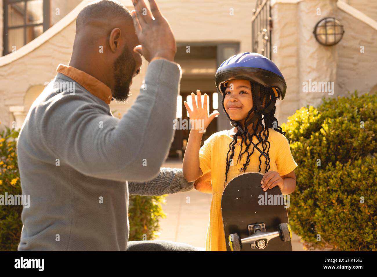 Sorridente ragazza afroamericana che indossa casco tenendo skateboard dando alto-cinque a padre in cantiere Foto Stock