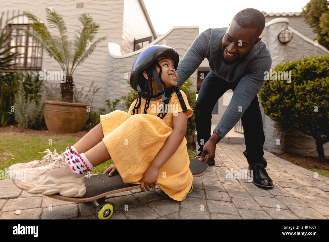 Felice padre africano americano che spinge la figlia sullo skateboard in cortile durante il fine settimana Foto Stock