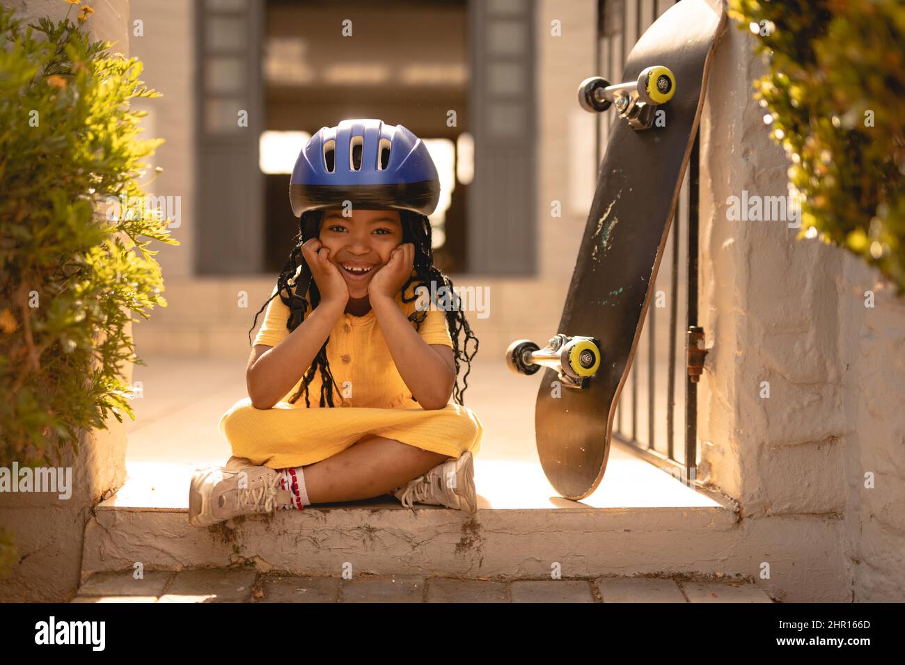 Ritratto di ragazza afro-americana allegra con casco seduto a gambe incrociate da skateboard al cancello Foto Stock