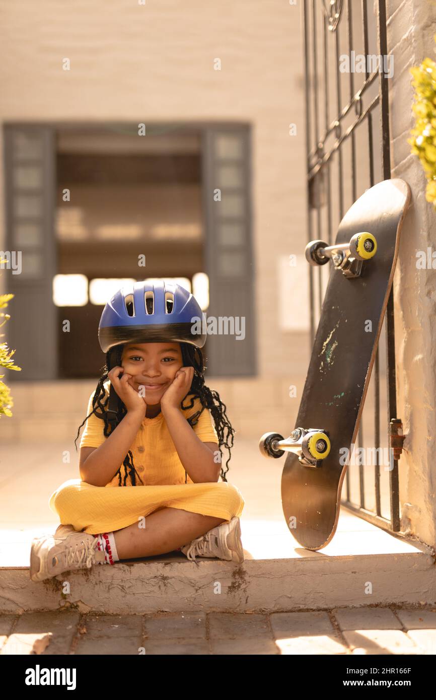 Ritratto di sorridente ragazza afroamericana con casco seduto a gambe incrociate da skateboard al cancello Foto Stock