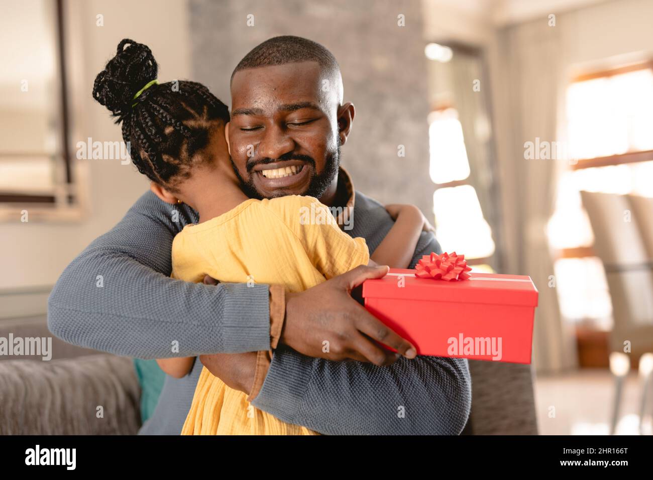 Felice padre afroamericano abbraccia la figlia dopo aver ricevuto il regalo rosso da lei a casa Foto Stock