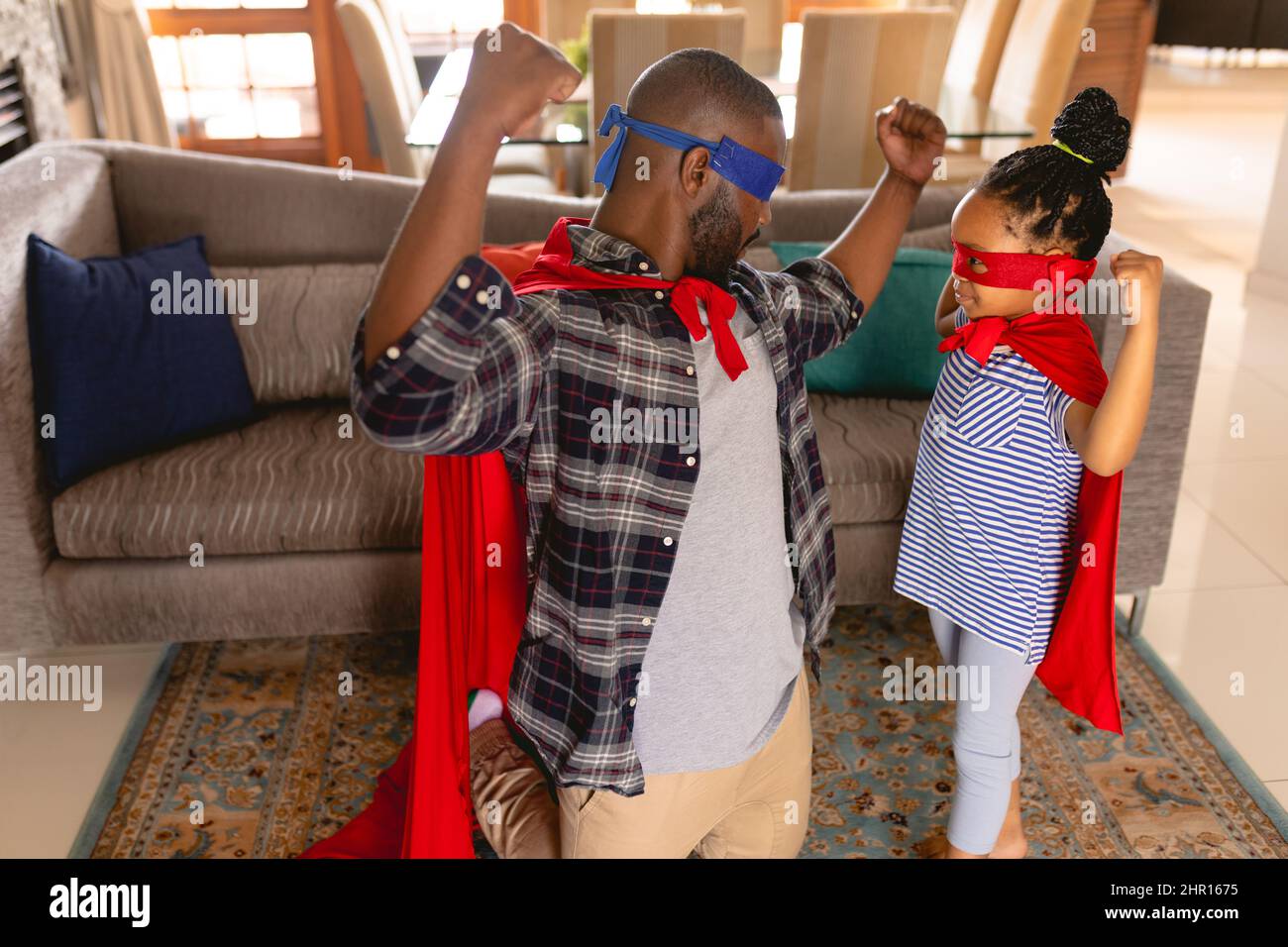 Il padre e la figlia afro-americana indossano costumi da supereroe che flettono i muscoli in salotto Foto Stock