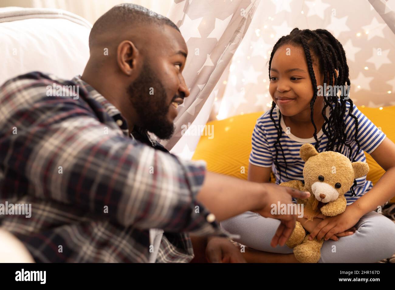 Sorridente ragazza afroamericana che tiene orsacchiotto guardando il padre che gioca con lei in tenda a casa Foto Stock