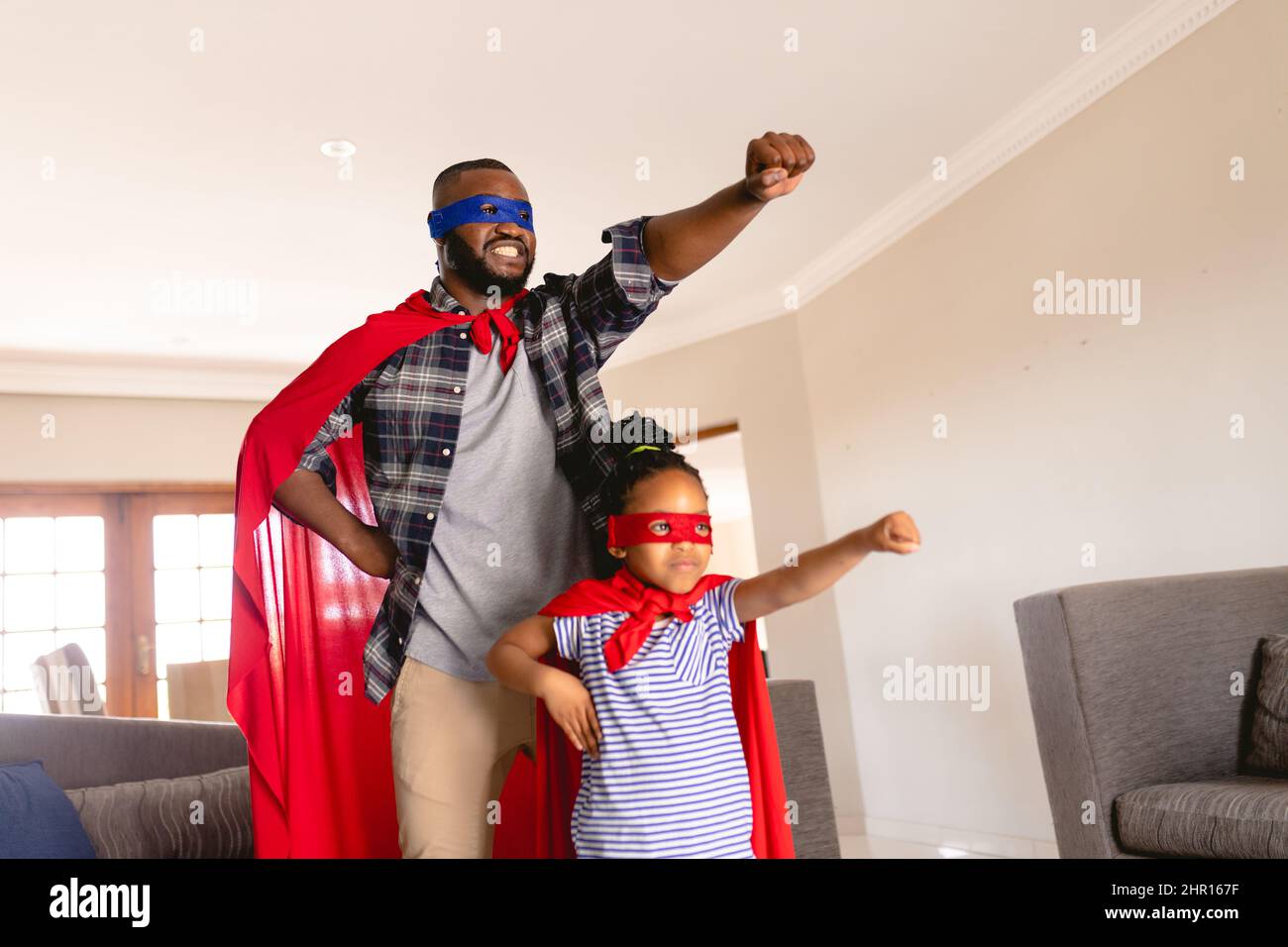 Sorridente padre e figlia afroamericana che indossa costumi da supereroe che giocano in salotto Foto Stock