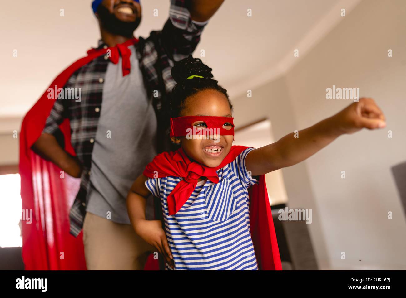 Ragazza allegra afroamericana con padre che indossa costumi da supereroe che giocano in salotto a casa Foto Stock