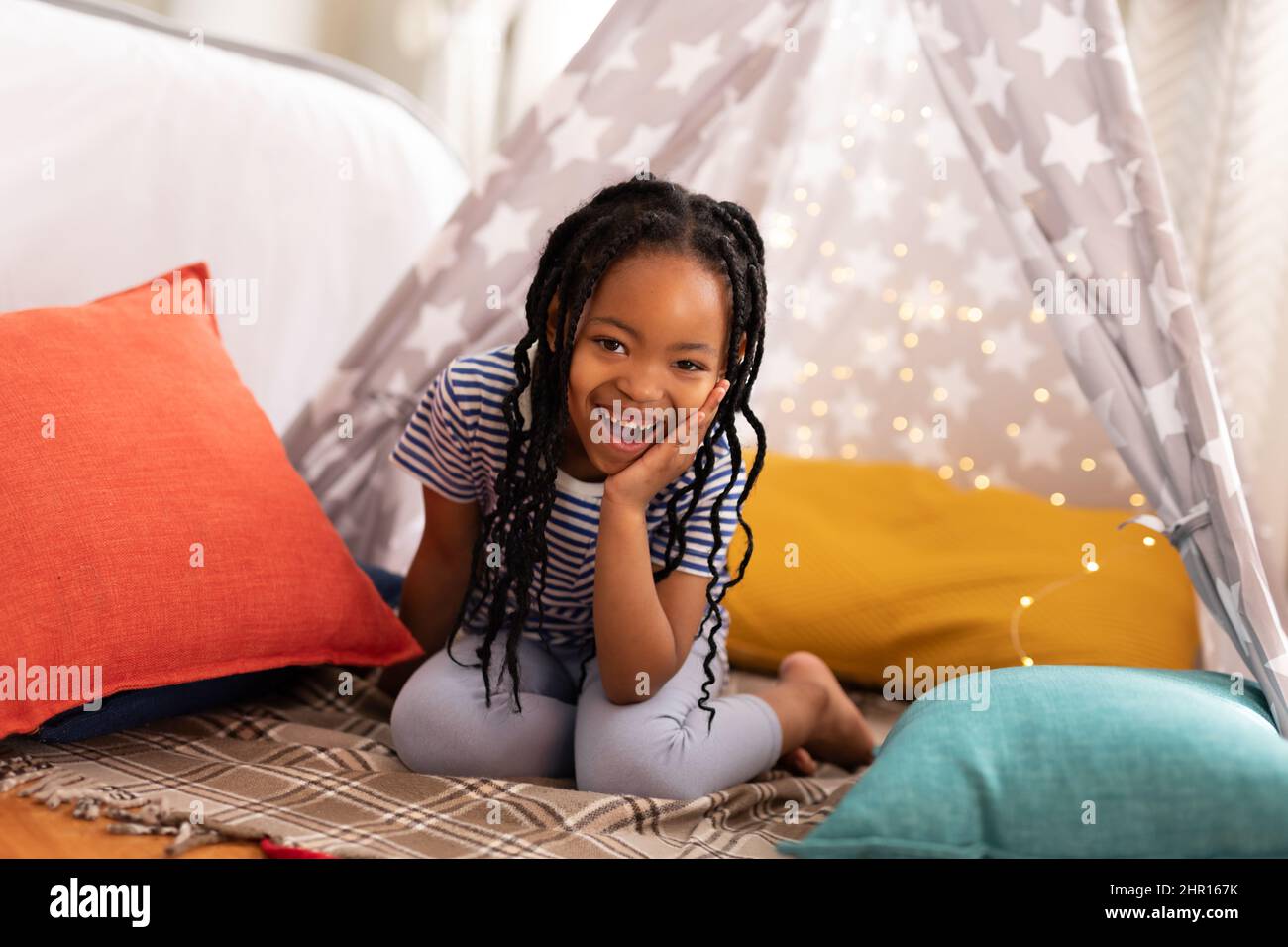 Ritratto di allegra ragazza afroamericana carina inginocchiarsi sul letto con tenda e cuscini a casa Foto Stock