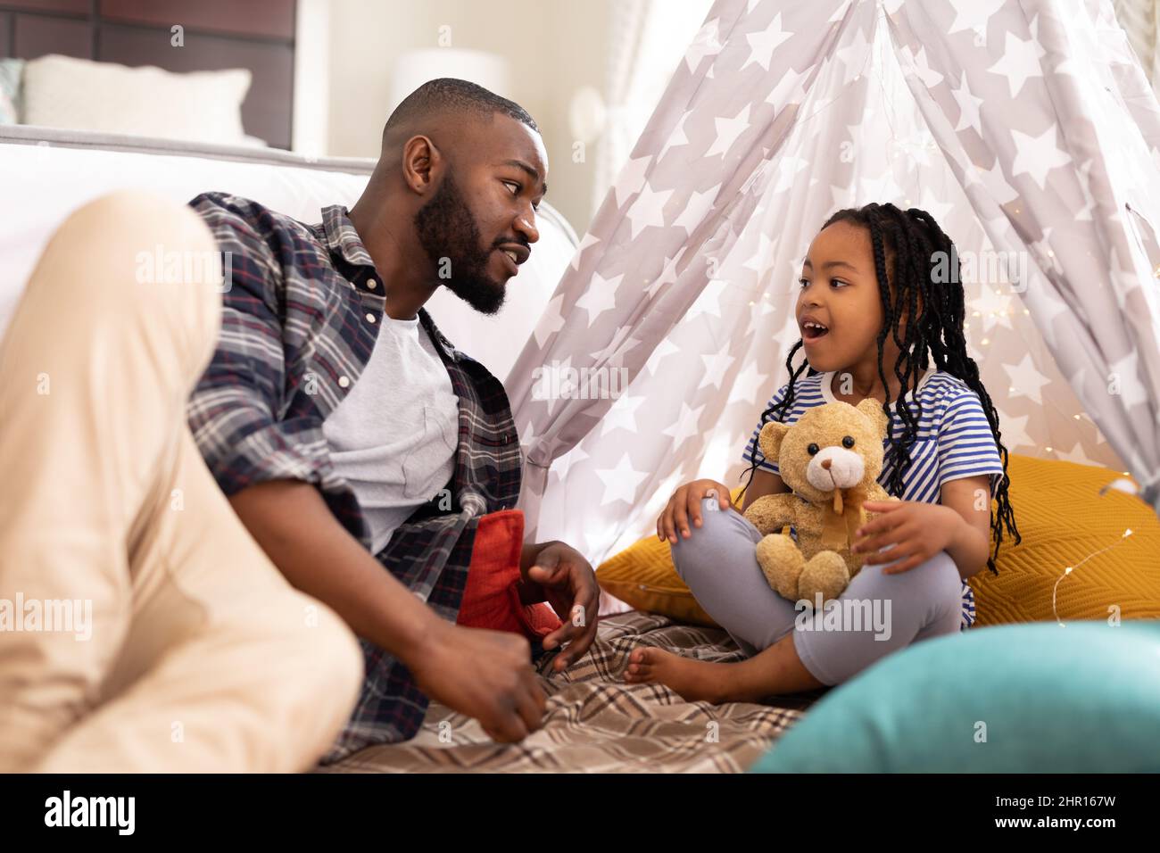 L'uomo afroamericano che parla alla figlia cute seduta con l'orso del teddy nella tenda in casa Foto Stock