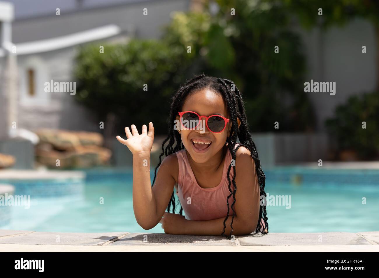 Ritratto della ragazza afro-americana felice che indossa occhiali da sole appoggiati a bordo piscina ondeggiando in giorno di sole Foto Stock