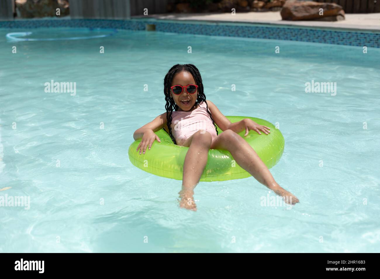 Ritratto di sorridente ragazza afroamericana con occhiali da sole seduti su anello gonfiabile in piscina Foto Stock