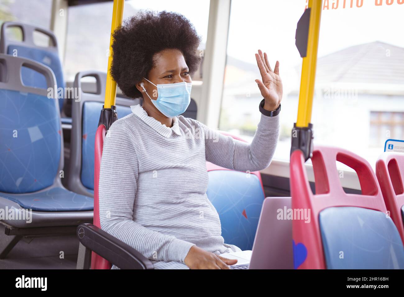 Donna d'affari afroamericana di metà adulto che indossa la maschera che ondita la mano mentre si siede con il laptop in autobus Foto Stock
