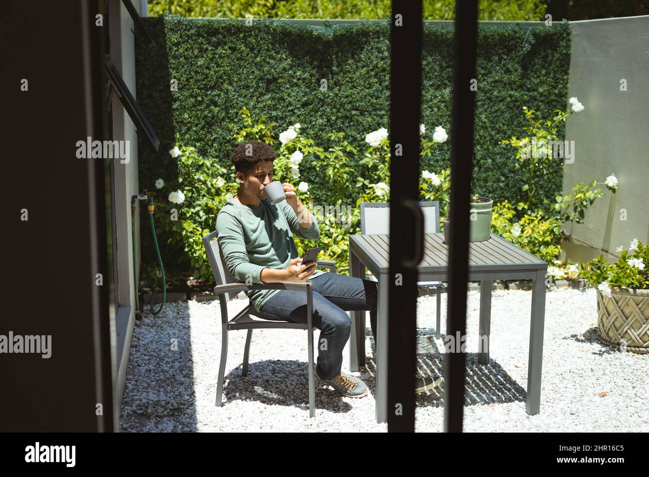 Uomo biraciale bere caffè mentre si siede su sedia in cortile in giorno di sole Foto Stock