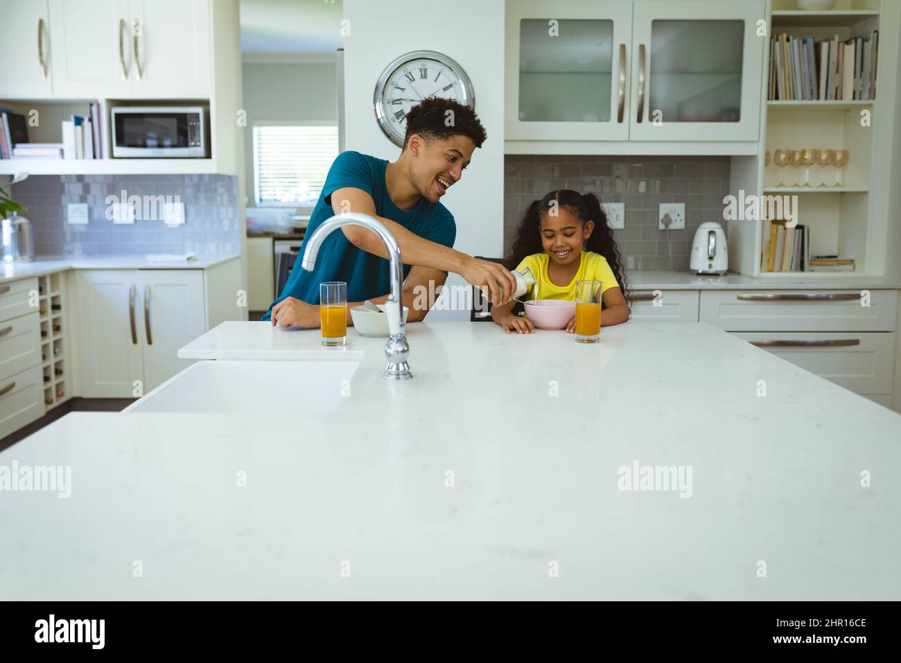 Felice padre multirazziale versando latte per la figlia seduta in cucina isola durante la prima colazione Foto Stock