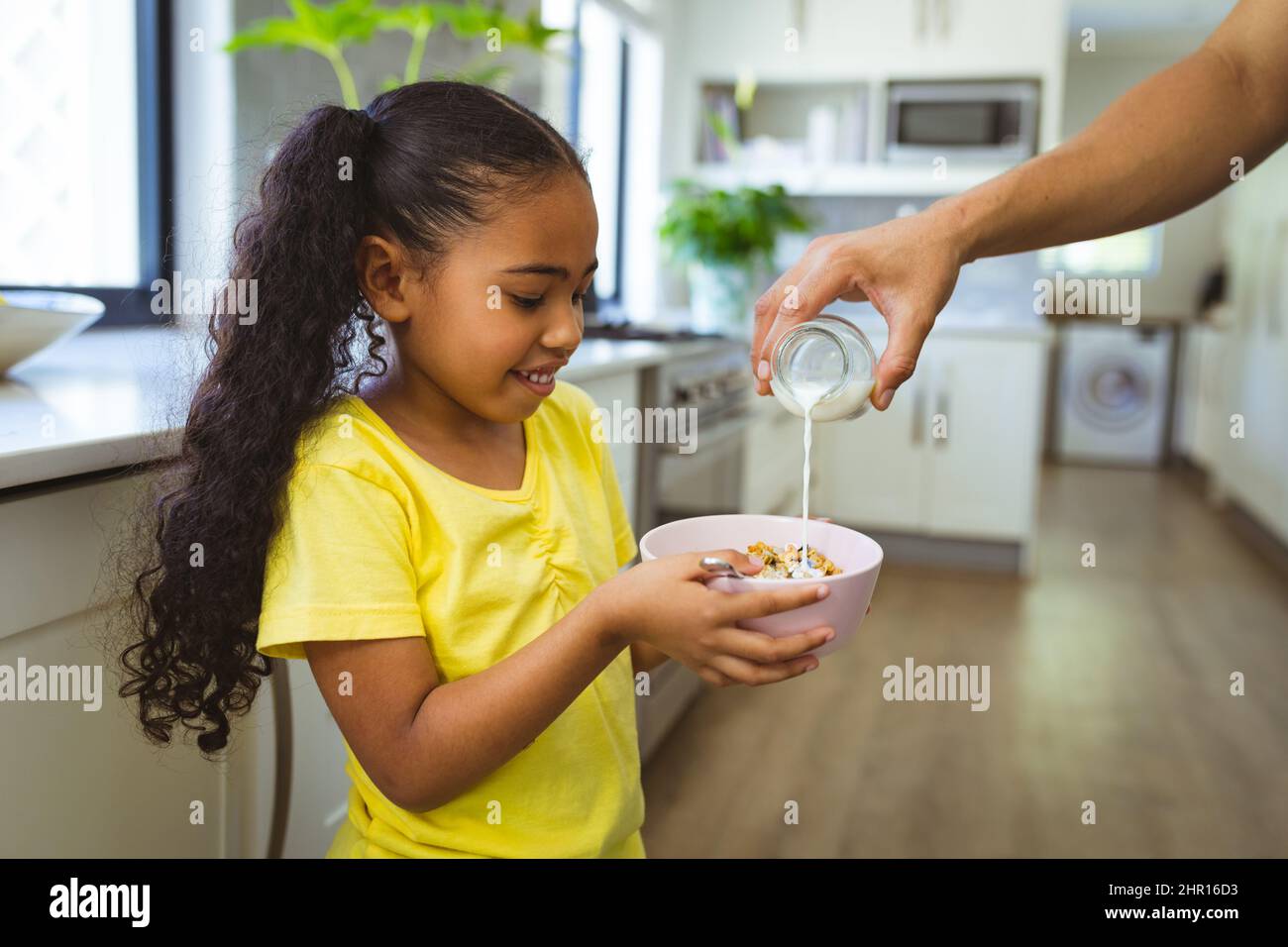 Mano di padre tagliata che versa il latte in ciotola di cereali per la figlia multirazziale in casa Foto Stock