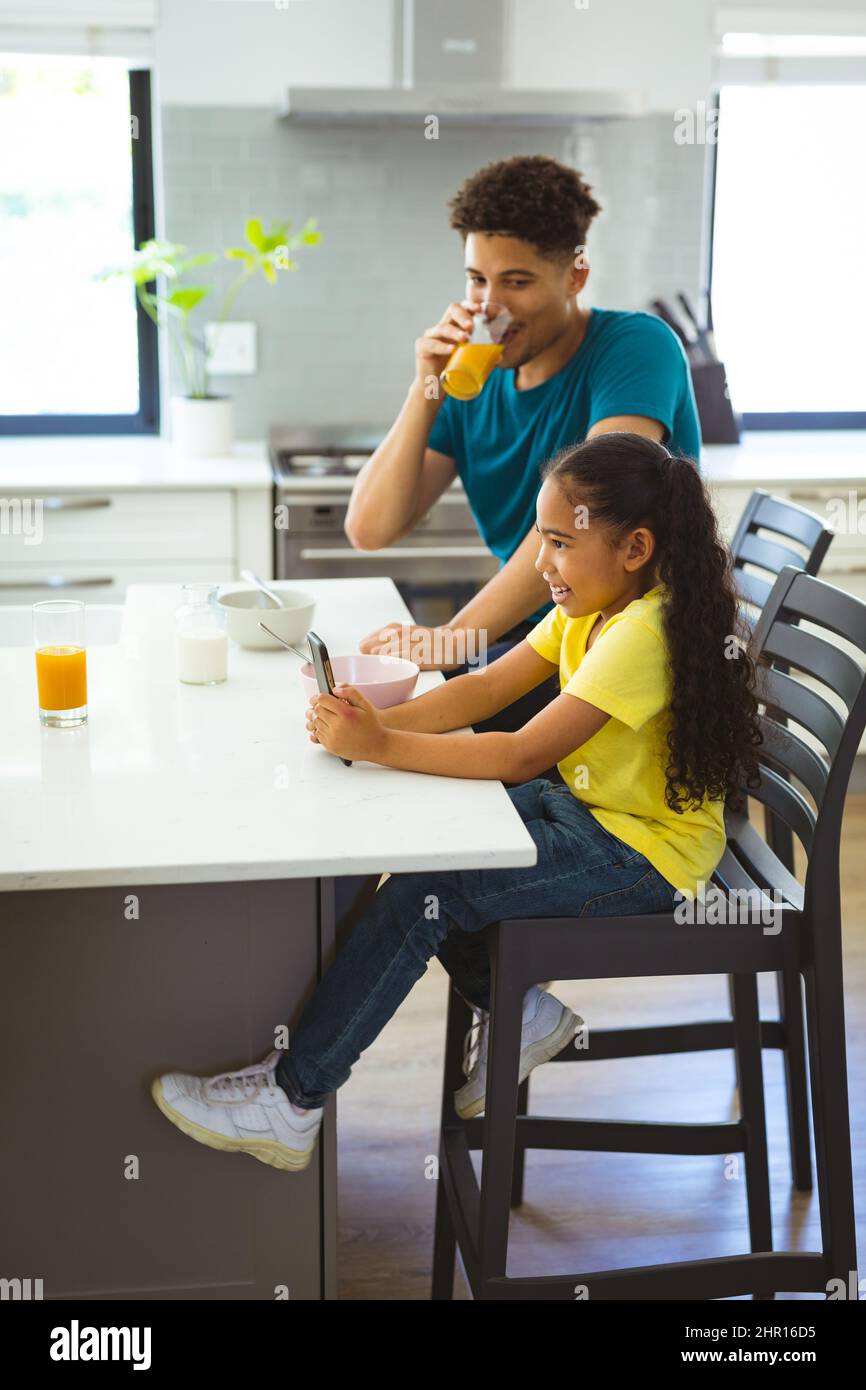 Multirazziale padre bere succo mentre si guarda la figlia seduta con smartphone in cucina Foto Stock