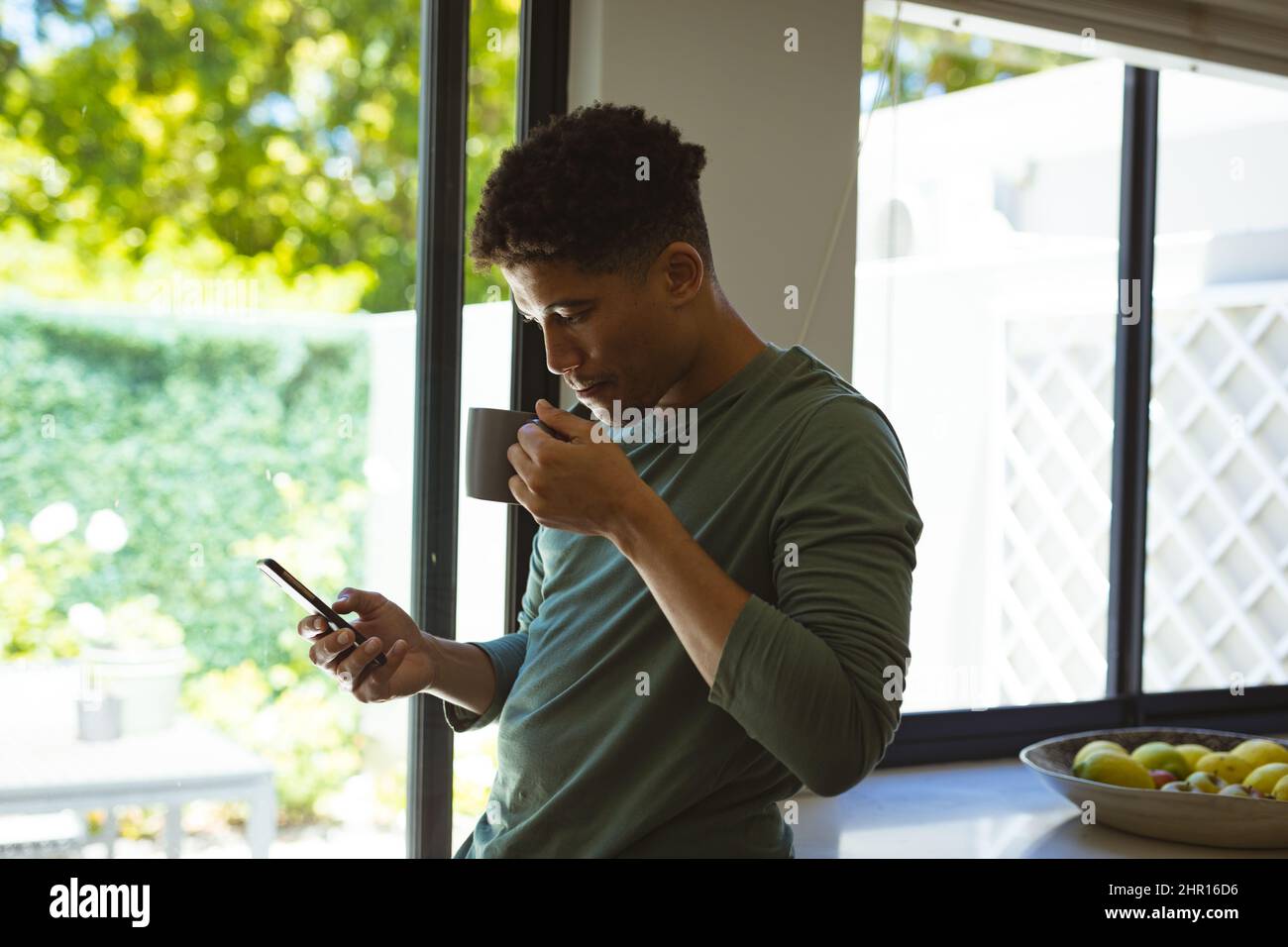 Giovane uomo biracale che beve caffè dalla tazza mentre usa lo smartphone in cucina a casa Foto Stock