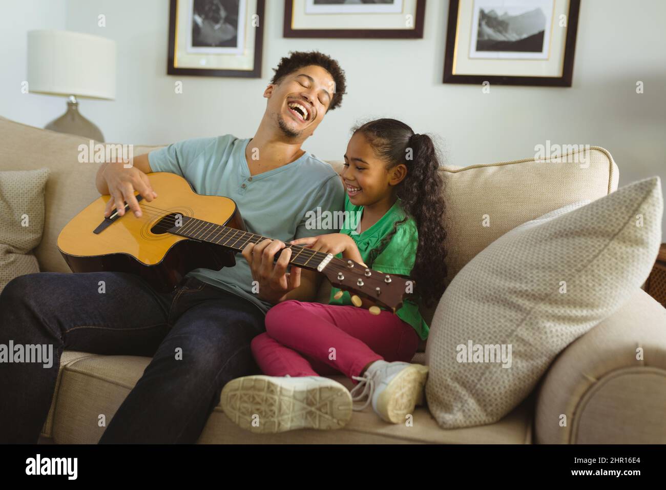 Allegro padre multirazziale che suona la chitarra mentre si siede con la figlia sul divano a casa Foto Stock