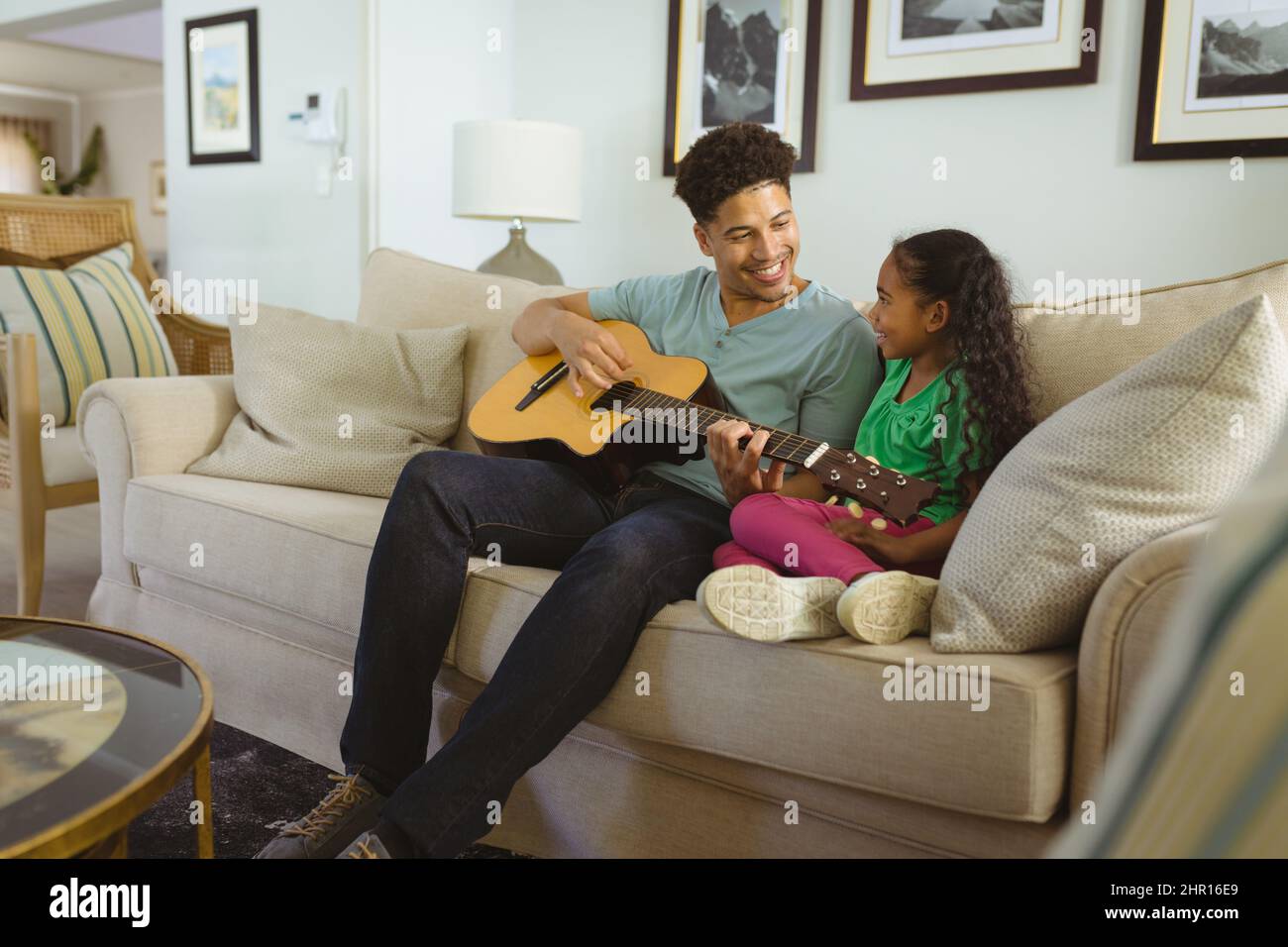 Felice padre multirazziale che suona la chitarra mentre si siede con la figlia sul divano in soggiorno Foto Stock