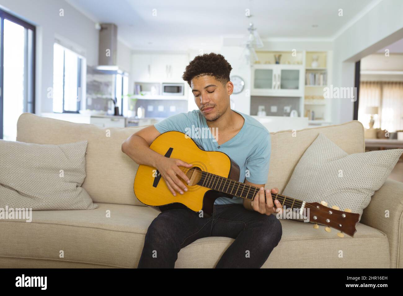 Giovane uomo biraciale che suona chitarra acustica mentre si siede sul divano nel soggiorno di casa Foto Stock