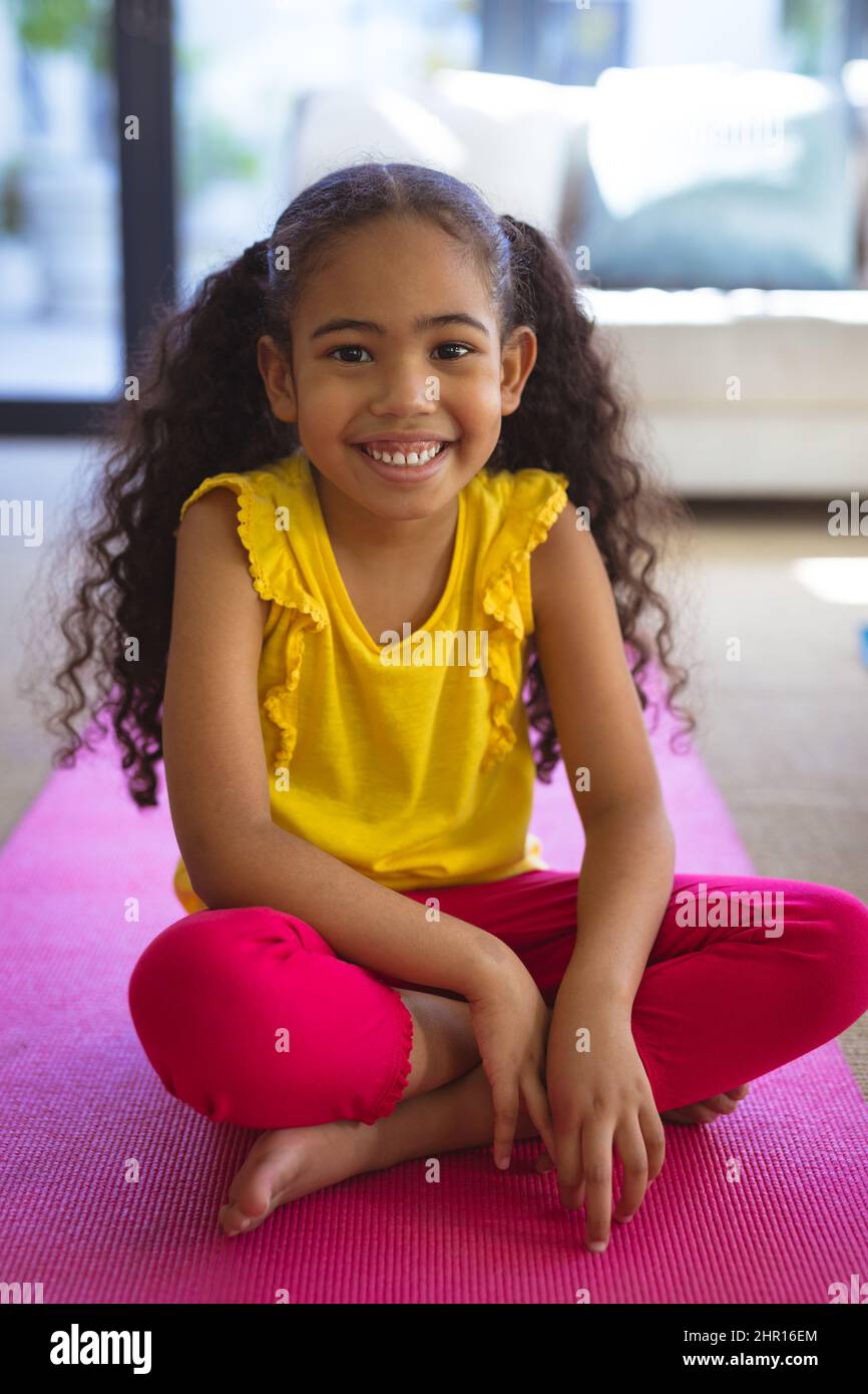 Ritratto a tutta lunghezza di sorridente ragazza biraciale seduta su tappeto rosa yoga in soggiorno a casa Foto Stock