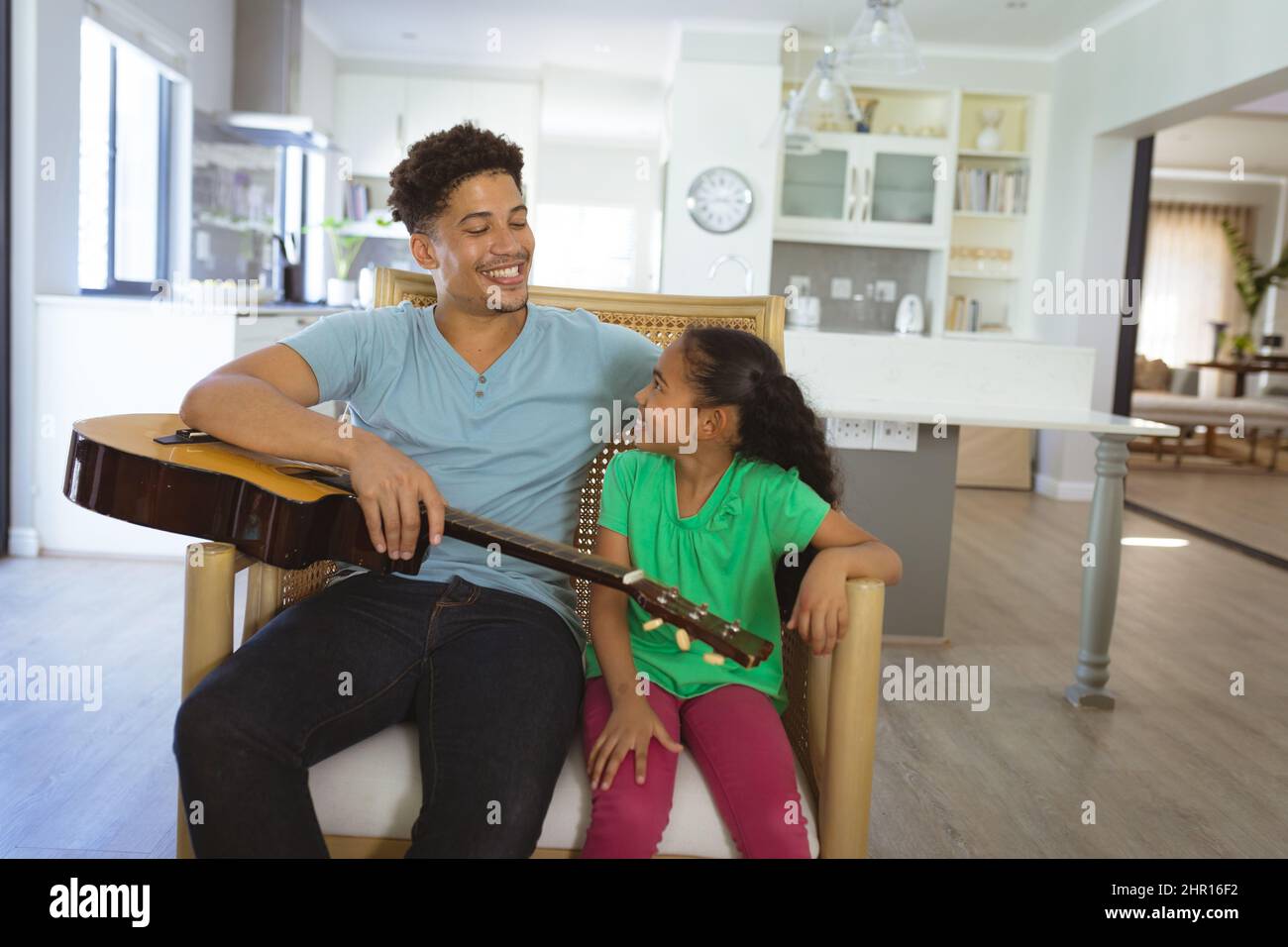 Sorridendo padre e figlia biraciale seduti insieme alla chitarra su poltrona nel soggiorno di casa Foto Stock