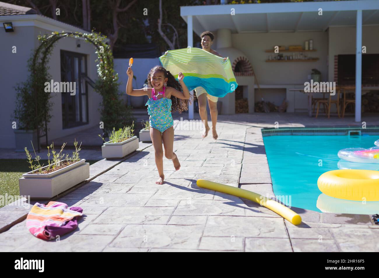 Giocosa ragazza biraciale che corre una testa di padre tenendo sarong a bordo piscina il giorno di sole Foto Stock