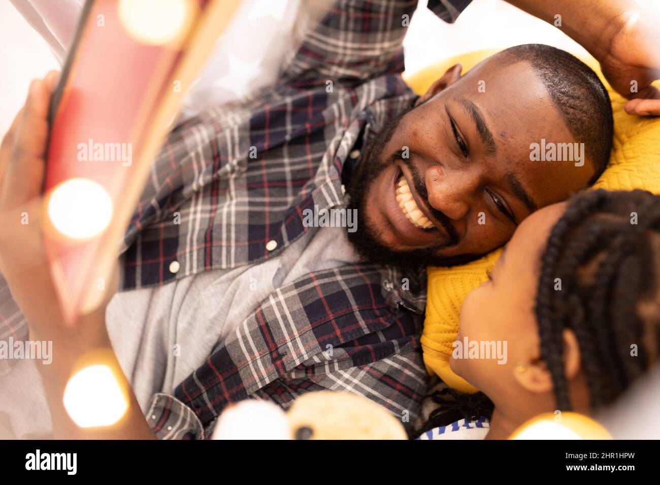 Allegro uomo afroamericano che legge storybook alla figlia mentre si trova insieme in tenda a casa Foto Stock
