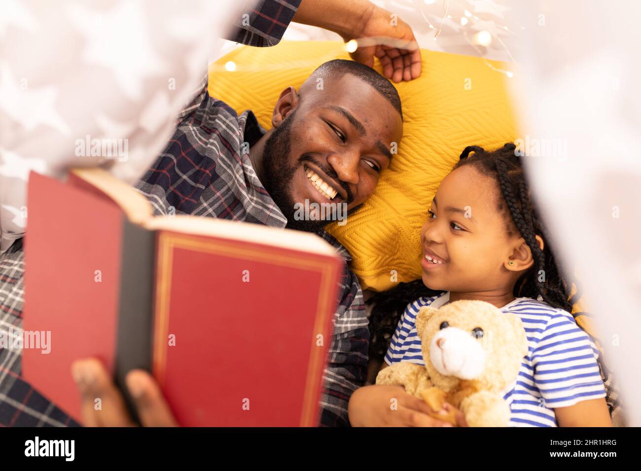 Sorridente uomo afroamericano che legge storybook alla figlia mentre si trova in tenda a casa Foto Stock