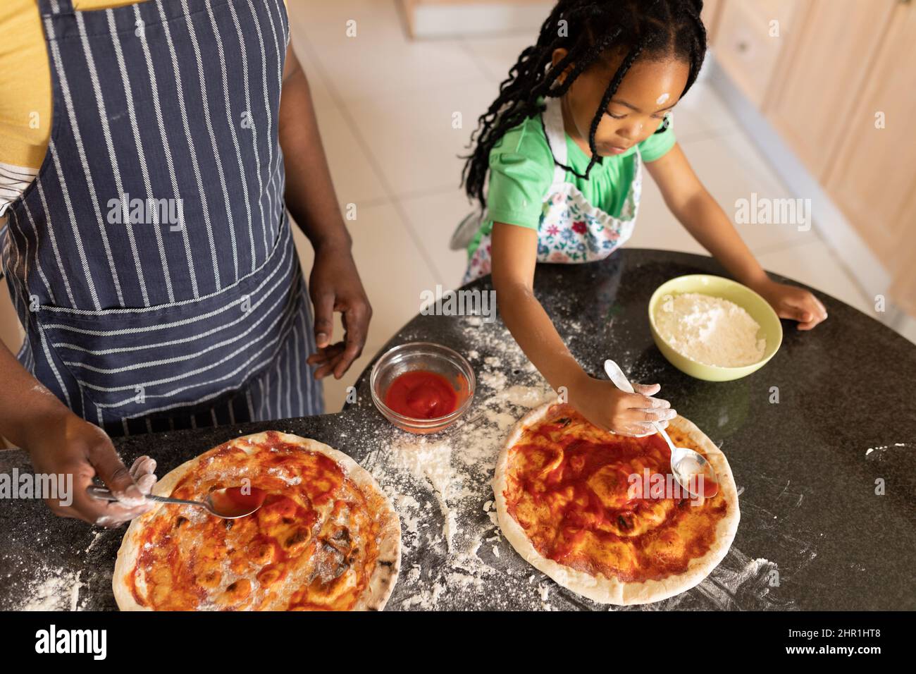 Ragazza africana americana che spalma la salsa sul pane della pizza dal padre che si alza al banco della cucina Foto Stock