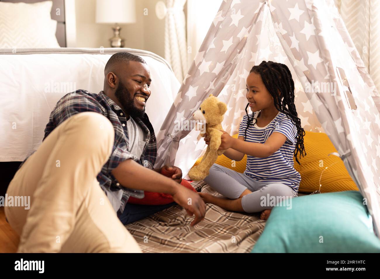 Felice padre e figlia afroamericana che giocano con orsacchiotto mentre si siede in tenda a casa Foto Stock