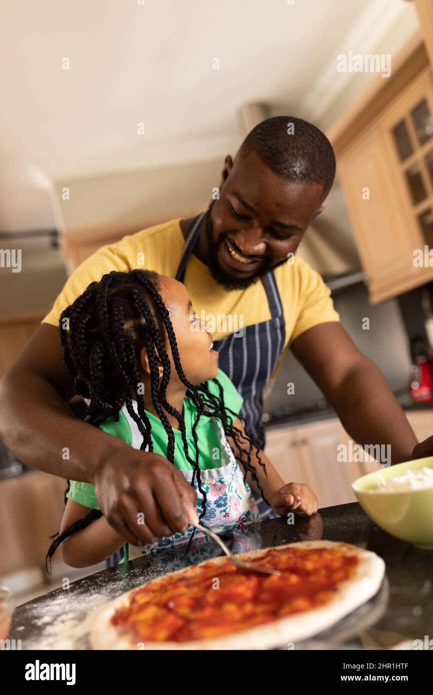 Sorridente padre afroamericano che aiuta la figlia a spalmare la salsa sulla pizza in cucina Foto Stock