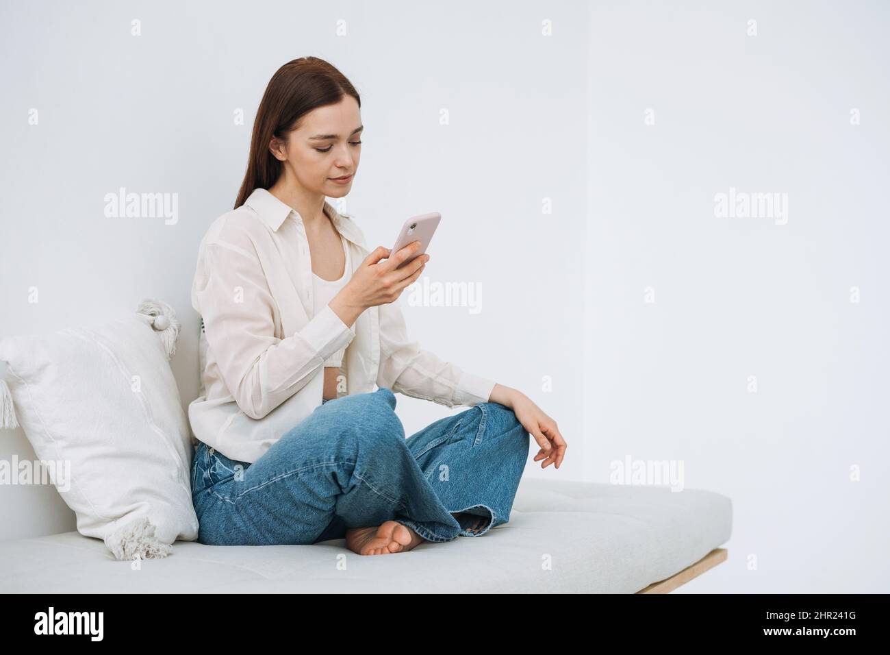 Bella donna sorridente adolescente studente ragazza con capelli lunghi e scuri in camicia bianca utilizzando il telefono cellulare in mano seduto sul divano a casa Foto Stock
