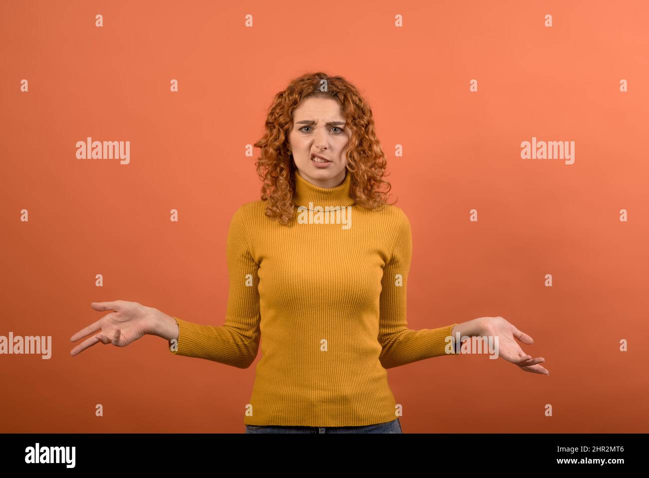 Giovane e attraente ragazza caucasica rossa in jumper arancione stendendo le braccia ai lati con espressione confusa isolato su studio arancione ba Foto Stock