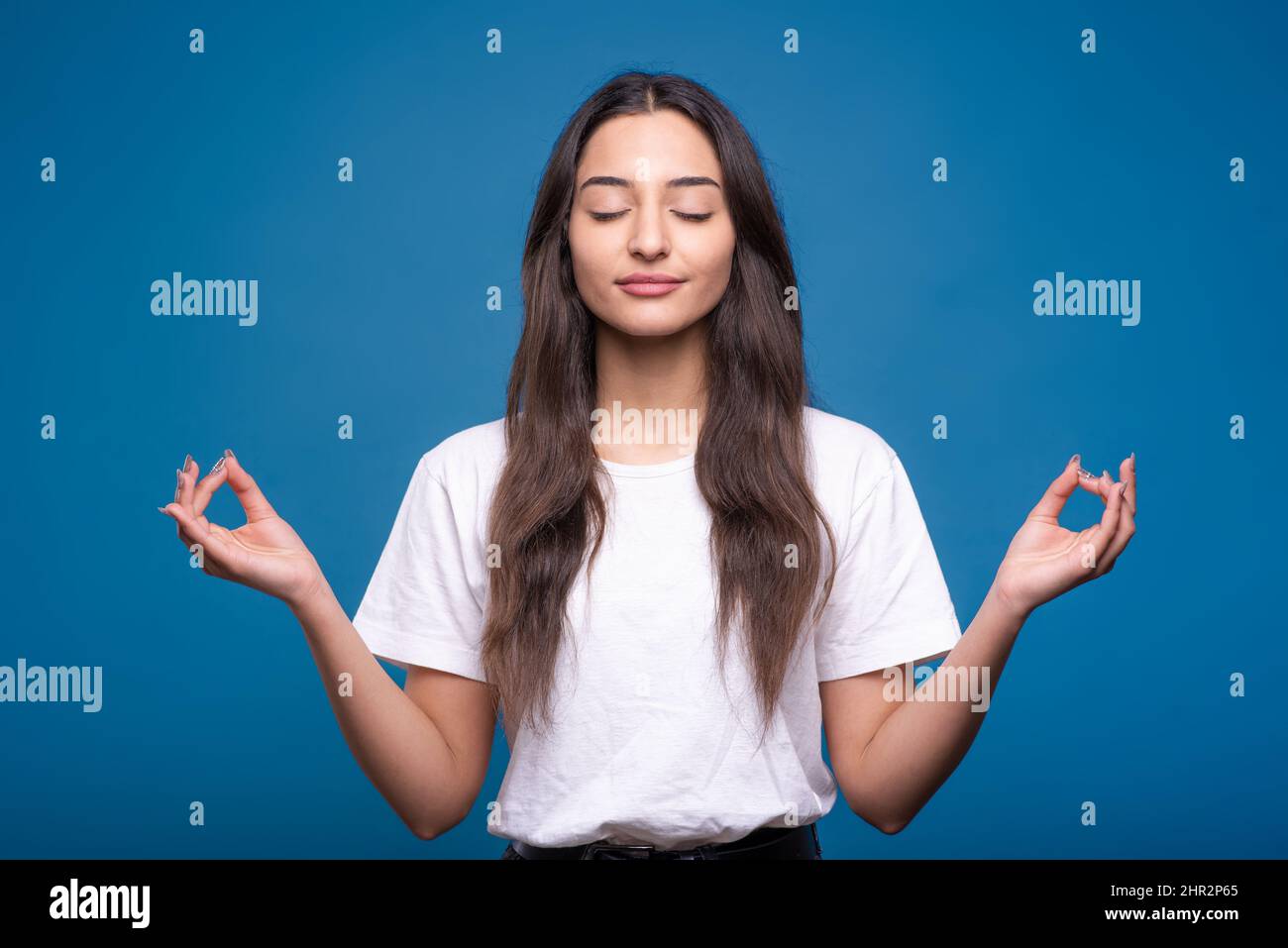 Bella ragazza bruna caucasica o araba in t-shirt bianca meditando in posizione lotus e mostrando il gesto zen isolato su sfondo blu studio. Foto Stock