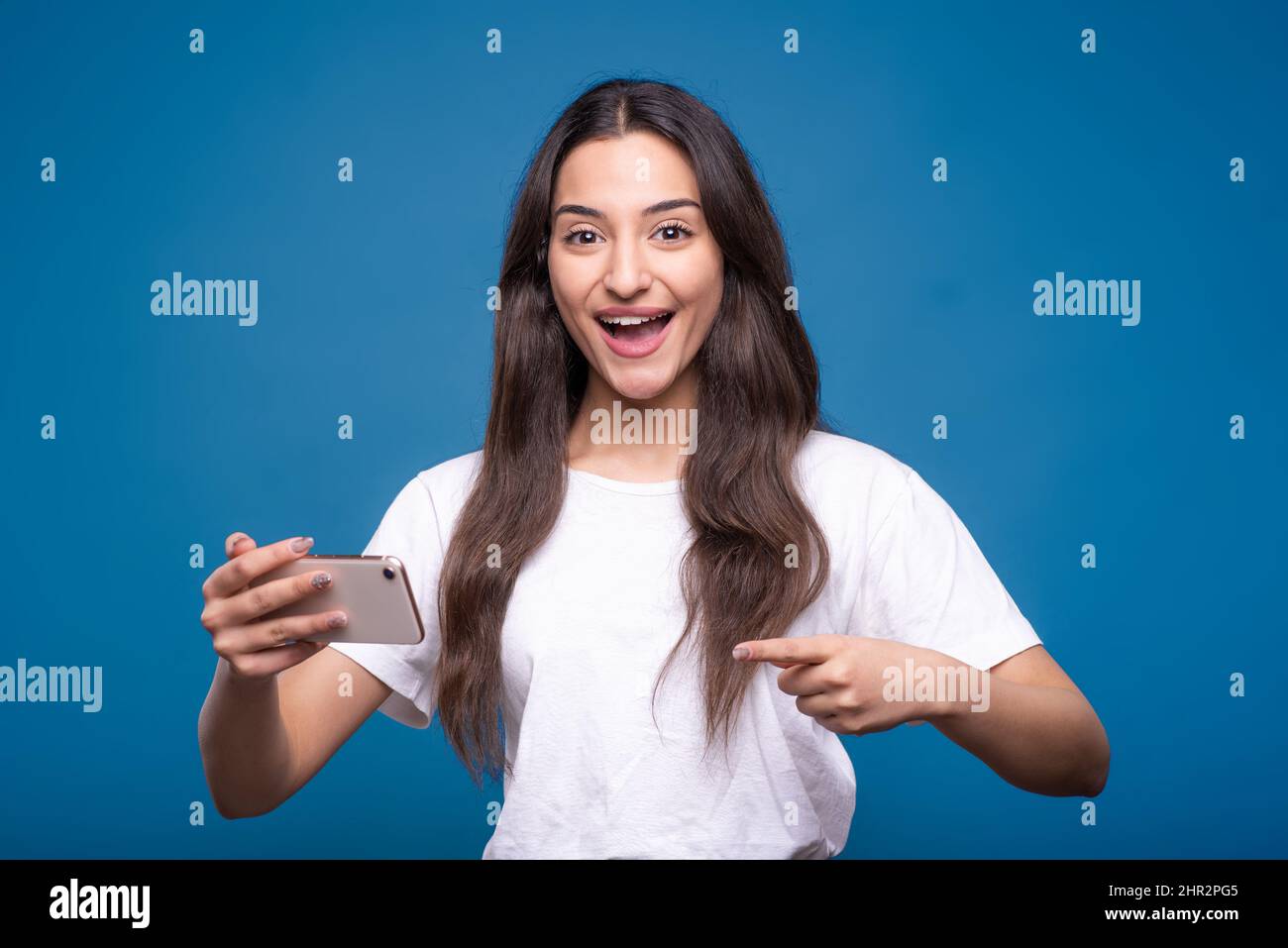 Bella ragazza bruna caucasica o araba in una t-shirt bianca è scioccata mentre si gioca su un telefono cellulare e puntando il dito su un'isola smartphone Foto Stock