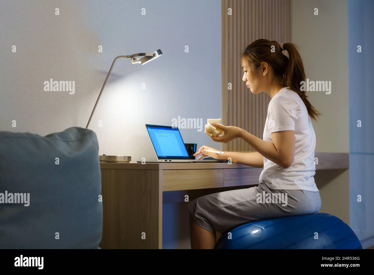Donna asiatica che si rilassa sulla palla yoga e mangia sandwich e chiacchiera online con il team di colleghi sul laptop mentre lavora da casa di notte. Foto Stock