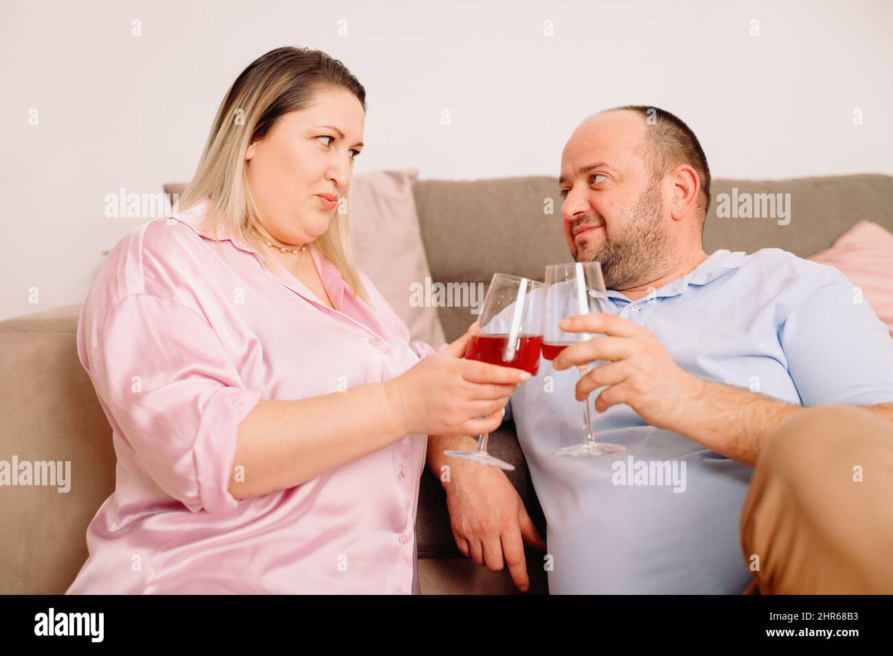 Felice coppia caucasica rilassarsi su un comodo divano a casa riposare tenendo in mano due bicchieri di vino e guardarsi con amore. Foto Stock