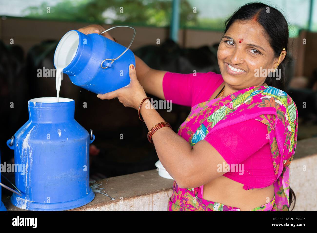 Felice sorridente donna indiana occupato di lavoro versando il latte nel contenitore mentre si guarda la macchina fotografica - concetto di produzione di latte, agri business, crescita e. Foto Stock