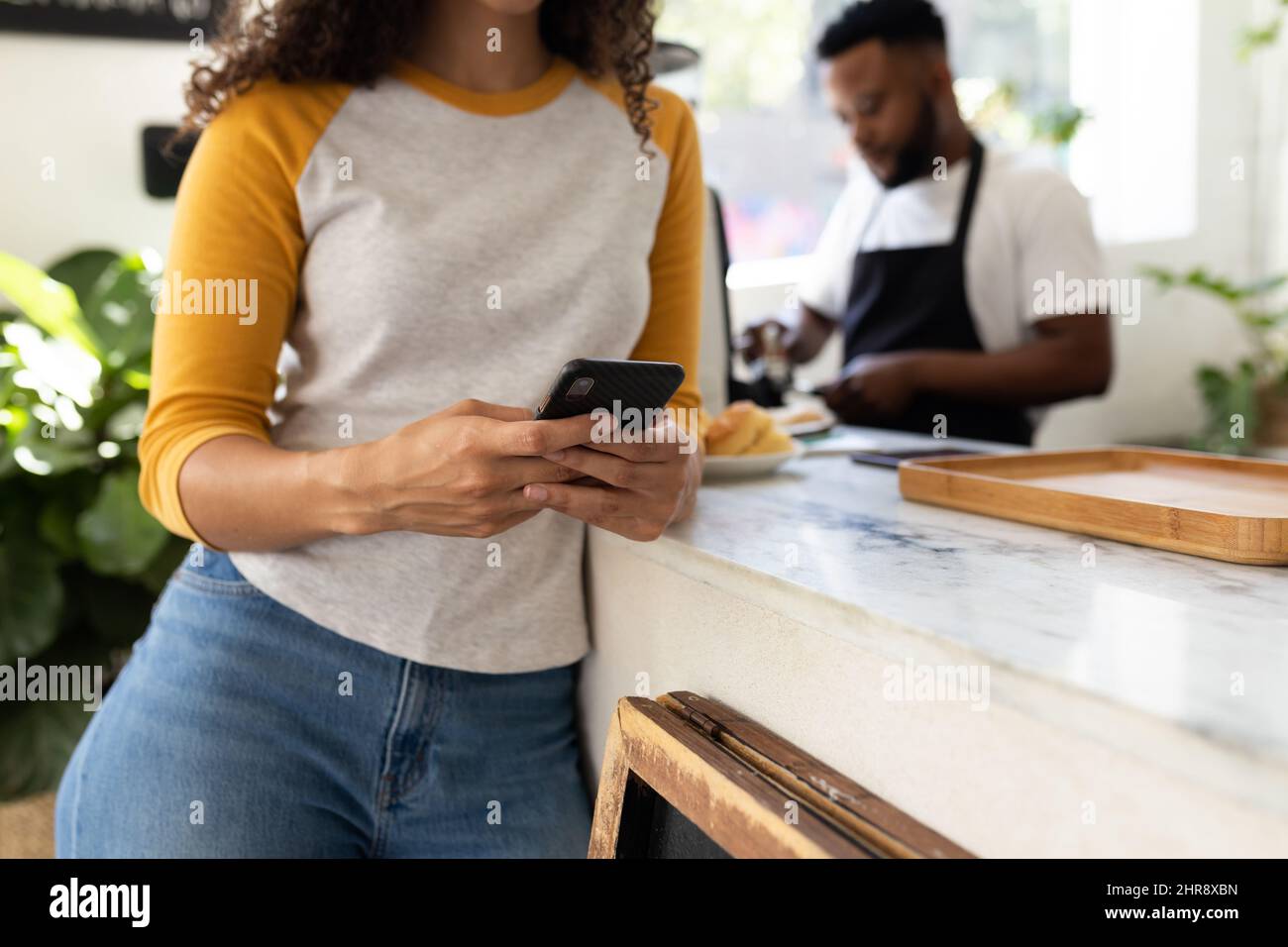 Metà della giovane donna afroamericana che usa lo smartphone al banco contro il barista in caffetteria Foto Stock