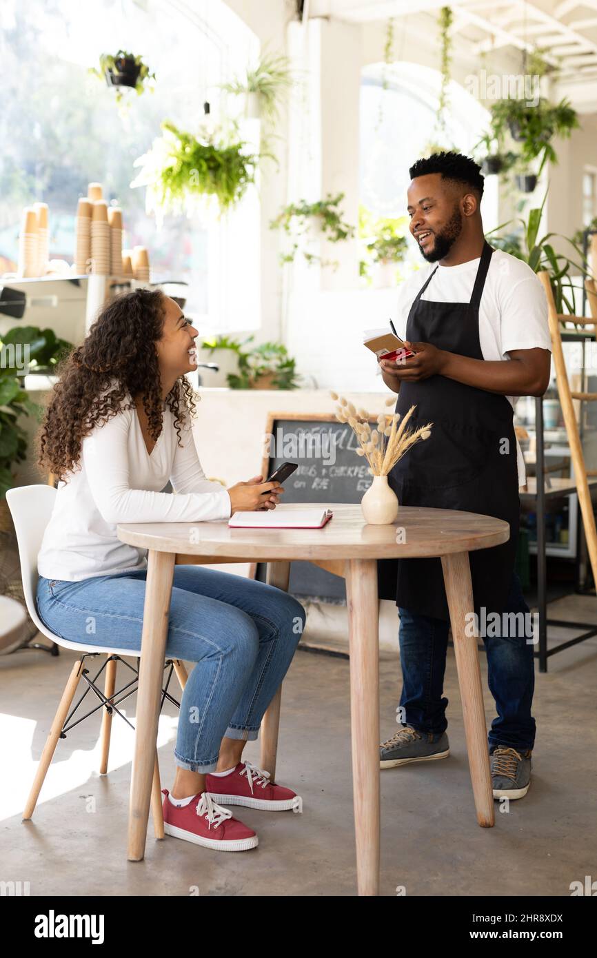 Sorridente barista afroamericano che prende ordine da giovane cliente femminile seduto in caffetteria Foto Stock